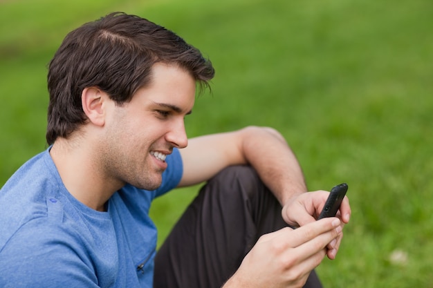 Junger Mann, der einen Text mit seinem Mobiltelefon beim Sitzen auf dem Gras sendet