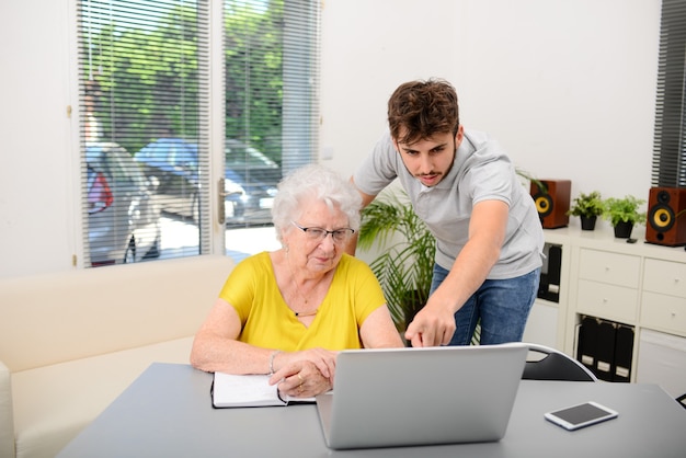 Junger Mann, der einer alten älteren Frau hilft, Papierkram und Verwaltungsverfahren mit einem Laptop zu Hause zu erledigen?