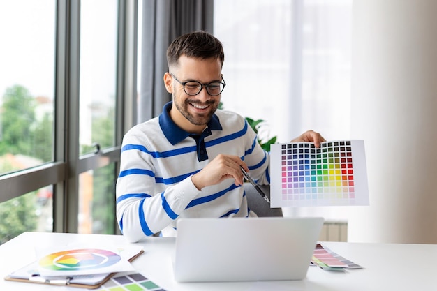 Junger mann, der farbmuster auf dem laptop-bildschirm im heimbüro zeigt und ein grafikdesignprojekt bei einem remote-meeting präsentiert millennial-designer, der gamma für die zukünftige hauseinrichtung auswählt