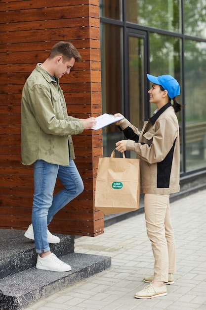 Junger Mann, der für Nahrungsmittellieferung im Dokument unterschreibt, während Frau ihn zu seinem Paket gibt, das sie draußen stehen
