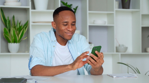 Junger Mann der gemischten Rasse, der am Tisch sitzt und SMS auf Smartphone liest Fokussierter tausendjähriger Geschäftsmann, der E-Mail in der mobilen Anwendung überprüft, die mit Partnern kommuniziert