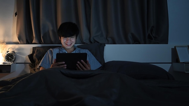 Junger Mann, der im Bett sitzt und nachts Filme anschaut oder mit einem digitalen Tablet im Internet surft.