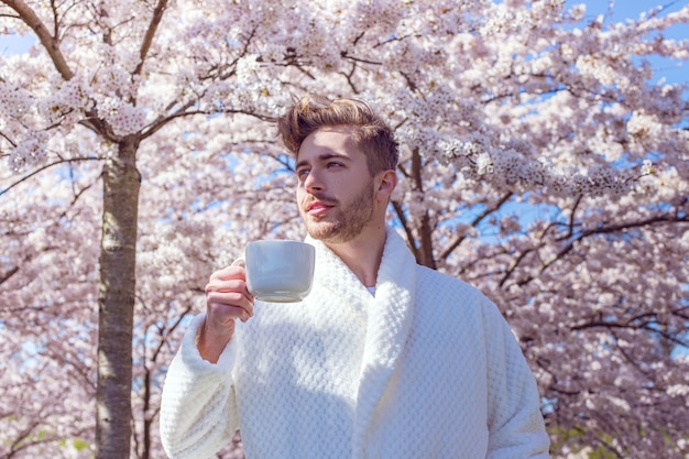 Junger Mann, der im blühenden Garten des Frühlings sich entspannt und Kaffee von der Tasse trinkt