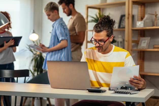 Junger Mann, der im modernen Büro arbeitet und Technologie nutzt, um seine Arbeit zu erledigen, und seine Kollegen stehen neben ihm und unterhalten sich