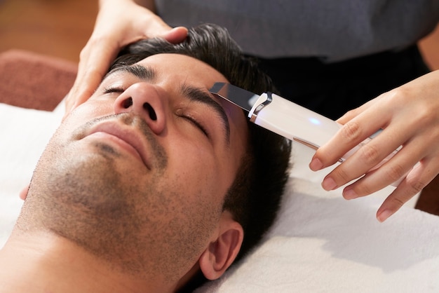 Junger Mann, der im Schönheitssalon eine Gesichtshautwäscherbehandlung mit Ultraschallspatel erhält