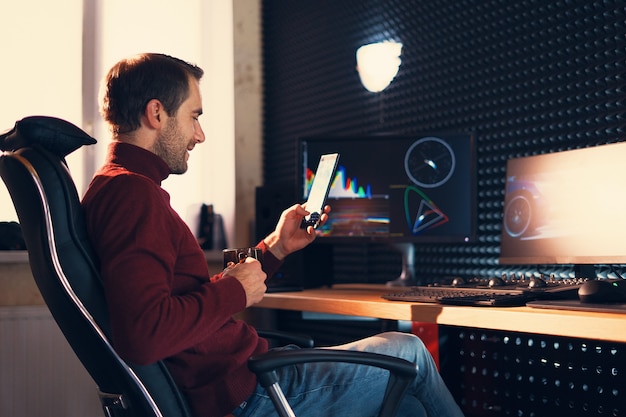 Junger Mann, der im Studio mit einem Smartphone und Computer arbeitet.