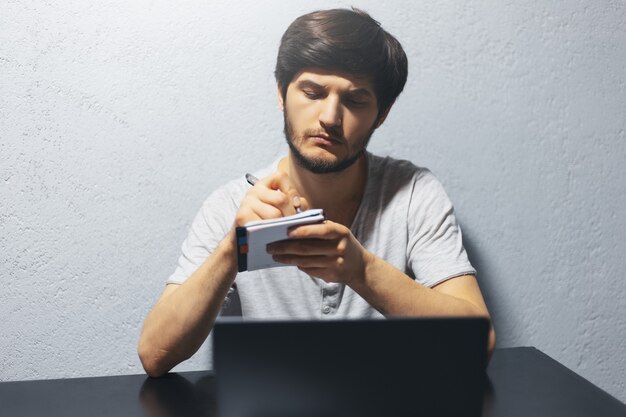 junger Mann, der in Notizbuchpapier schreibt. Arbeiten am Laptop, am schwarzen Tisch.