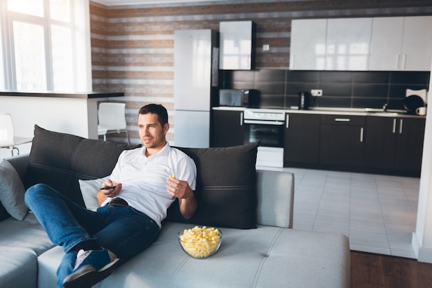 Junger Mann, der in seiner eigenen Wohnung fernsieht. Setz dich alleine auf die Couch und iss Snacks. Verwenden Sie die Fernbedienung zum Umschalten der Fernsehkanäle.