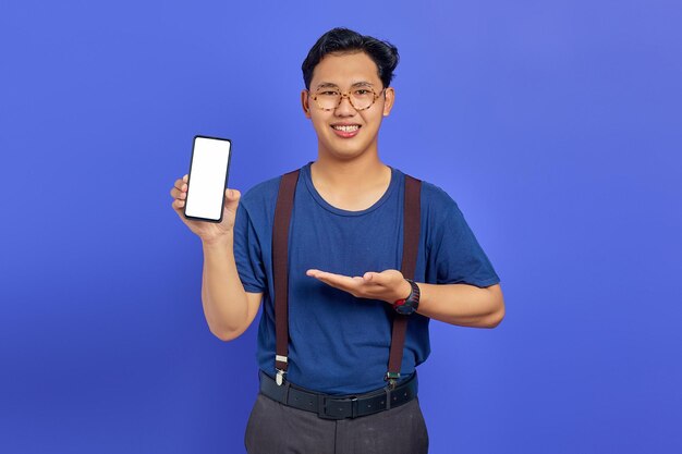 Junger Mann, der lächelt, während er Produkt präsentiert und Smartphone mit Handfläche auf violettem Hintergrund zeigt
