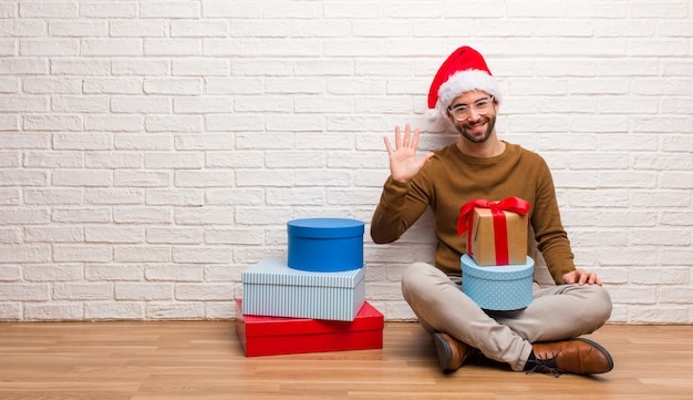 Junger Mann, der mit den Geschenken feiern das Weihnachten zeigt Nr. Fünf sitzt