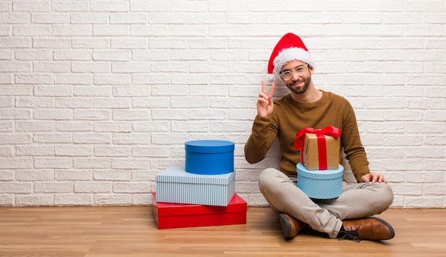 Junger Mann, der mit den Geschenken feiern das Weihnachten zeigt Nummer zwei sitzt