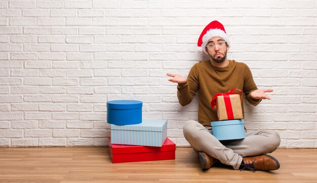 Junger Mann, der mit den Geschenken feiern Weihnachten verwirrt und zweifelhaft sitzt