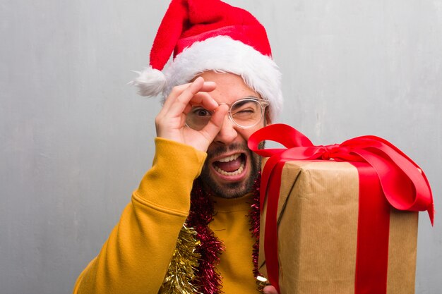 Junger Mann, der mit den Geschenken feiert Weihnachten sitzt, die Geste eines Fernglases machen