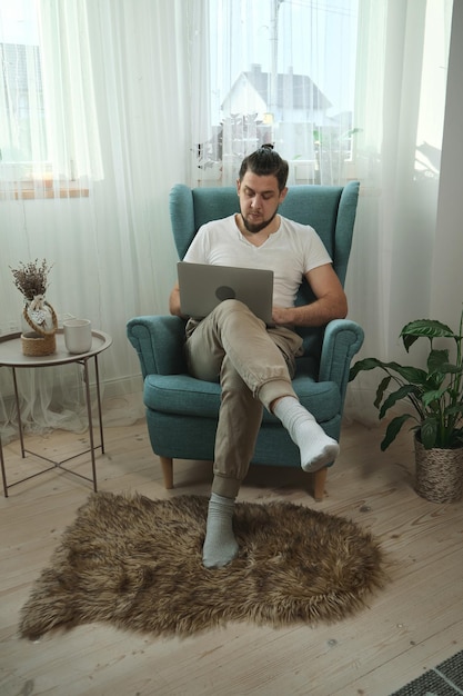 Junger Mann, der mit Laptop tippt und zu Hause auf dem Sessel sitzt