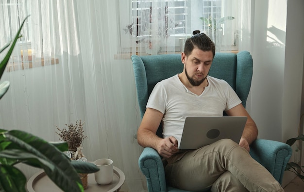 Junger Mann, der mit Laptop tippt und zu Hause auf dem Sessel sitzt