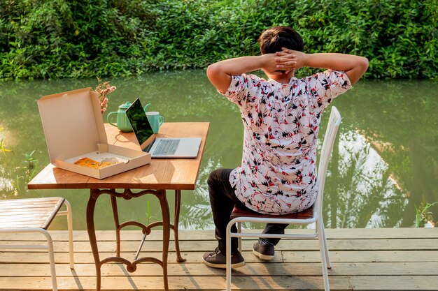 Junger Mann, der mit Pizza auf Tisch und Laptop entspannt