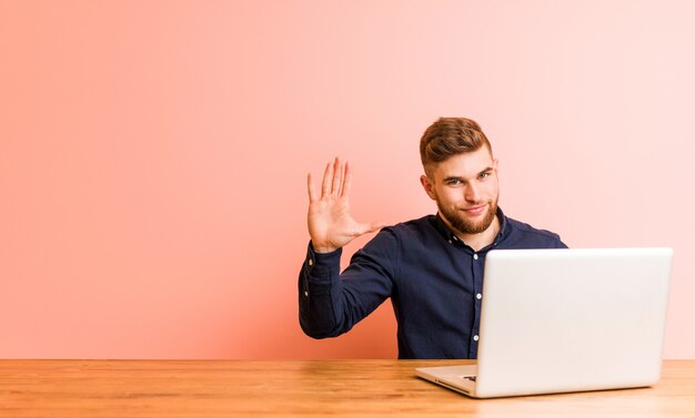 Junger Mann, der mit seinem Laptoplächeln freundlich arbeitet, Nr. Fünf mit den Fingern zeigend.