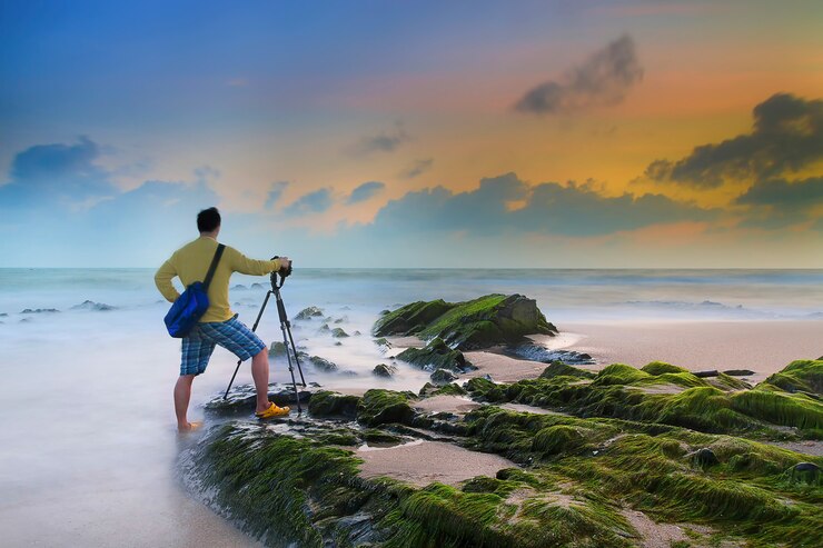 Junger Mann, der mit Stativ am Strand nach Sonnenuntergang fotografiert