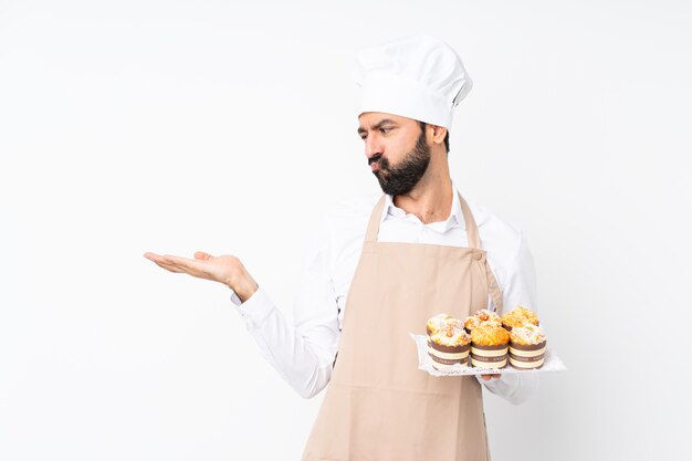 Junger Mann, der Muffinkuchen über lokalisiertem weißem haltenem copyspace mit Zweifeln hält