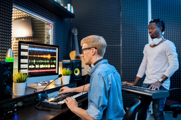 Junger Mann, der Musik durch Computer aufzeichnet, während afrikanischer Kerl Klavier im Studio der Aufzeichnungen spielt