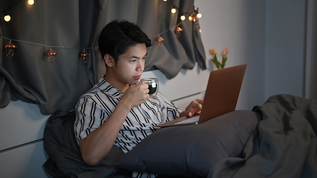 Junger Mann, der nachts heißen Tee und Laptop auf dem Bett trinkt.