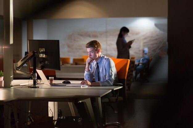 Junger Mann, der nachts im dunklen Büro am Computer arbeitet. Der Designer arbeitet in der späteren Zeit.