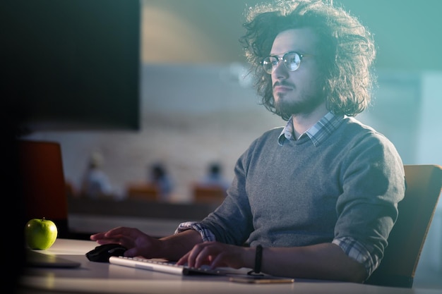 Junger Mann, der nachts im dunklen Büro am Computer arbeitet. Der Designer arbeitet in der späteren Zeit.