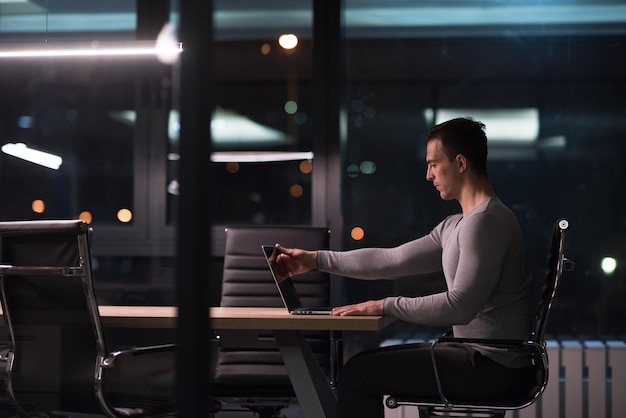 Junger Mann, der nachts im dunklen Büro am Laptop arbeitet. Der Designer arbeitet in der späteren Zeit.