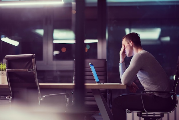 Junger Mann, der nachts im dunklen Büro am Laptop arbeitet. Der Designer arbeitet in der späteren Zeit.