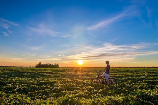 Junger Mann, der nahes Fahrrad im Morgensonnenaufgang mit wunderbaren Strahlen und Morgennebel während des aktiven Tages des ruhigen Sommers steht