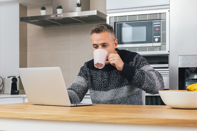 Junger Mann, der Notizbuch beim Frühstück, Lesen, Schreiben von E-Mail oder Online-Sitzen in der modernen Küche verwendet