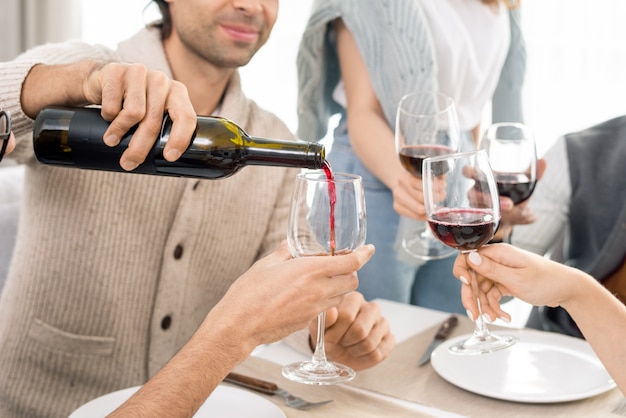 Junger Mann, der Rotwein von Flasche in Weingläser seiner Freunde gießt, während Feiertag durch gedienten Tisch zusammen feiert