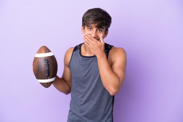 Junger Mann, der Rugby spielt, isoliert auf violettem Hintergrund, glücklich und lächelnd, den Mund mit der Hand bedeckend