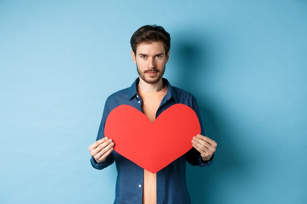 Junger Mann, der Seelenverwandten am Valentinstag sucht, großes rotes Herz hält und Kamera betrachtet, über blauem Hintergrund stehend.