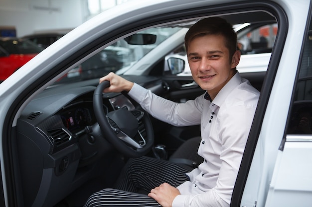 Junger Mann, der sich für ein neues Auto zum Kauf im Autohaussalon entscheidet und in einem neuen Auto sitzt