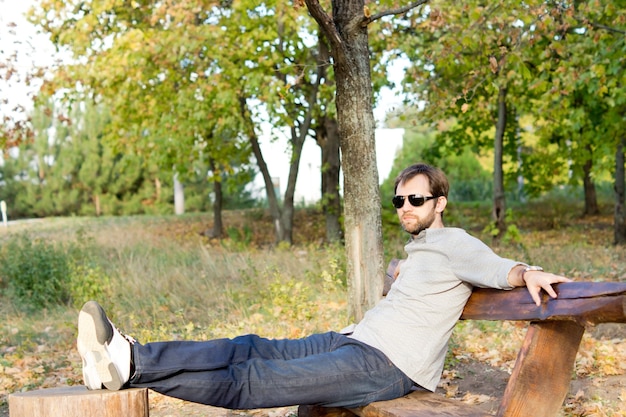 Junger Mann, der sich in der Sonne entspannt, die auf einer rustikalen Holzbank mit seinen Füßen oben auf einem Baumstumpf sitzt