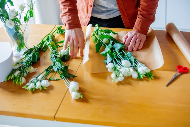 Junger Mann, der weiße Rosen auf einem Tisch in der Küche einwickelt. Eigenes Geschäftskonzept