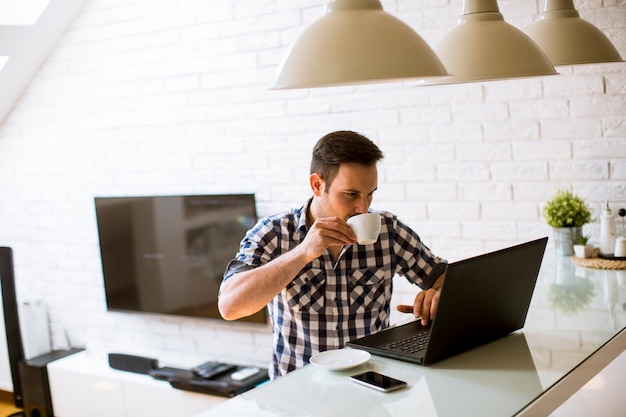 Junger Mann, der zu Hause auf Küchenschreibtisch sitzt und Laptop und Kaffee trinkt