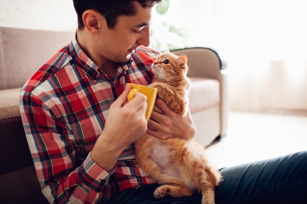 Junger Mann, der zu Hause eine Katze hält und Tee trinkt