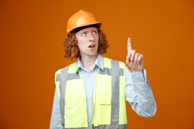 Junger mann des baumeisters in bauuniform und schutzhelm, der besorgt nach oben schaut und mit dem zeigefinger nach oben zeigt, der über orangefarbenem hintergrund steht