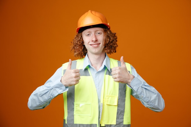 Junger Mann des Baumeisters in Bauuniform und Schutzhelm, der glücklich und positiv lächelnd in die Kamera blickt und fröhlich die Daumen nach oben zeigt, die über orangefarbenem Hintergrund stehen