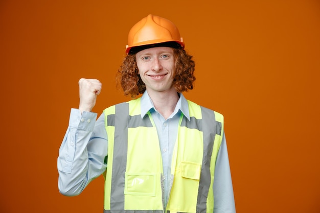 Junger Mann des Baumeisters in Bauuniform und Schutzhelm, der glücklich und positiv lächelnd in die Kamera blickt und mit dem Daumen nach hinten zeigt, der über orangefarbenem Hintergrund steht