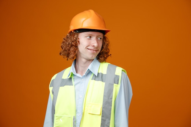 Junger Mann des Baumeisters in Bauuniform und Schutzhelm, der mit einem Lächeln auf dem glücklichen Gesicht, das über orangefarbenem Hintergrund steht, beiseite schaut