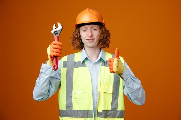 Junger Mann des Baumeisters in Bauuniform und Schutzhelm mit Schraubenschlüssel, der in die Kamera blickt und fröhlich lächelt und den Daumen nach oben zeigt, der über orangefarbenem Hintergrund steht