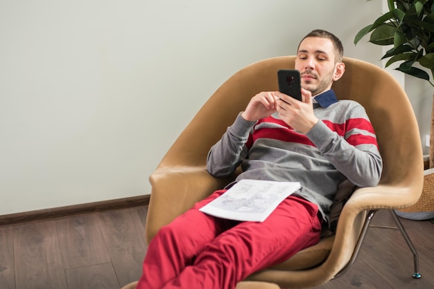 Junger Mann des Geschäfts, der sein intelligentes Telefon beim Lügen auf der Couch zu Hause oder im Büro betrachtet