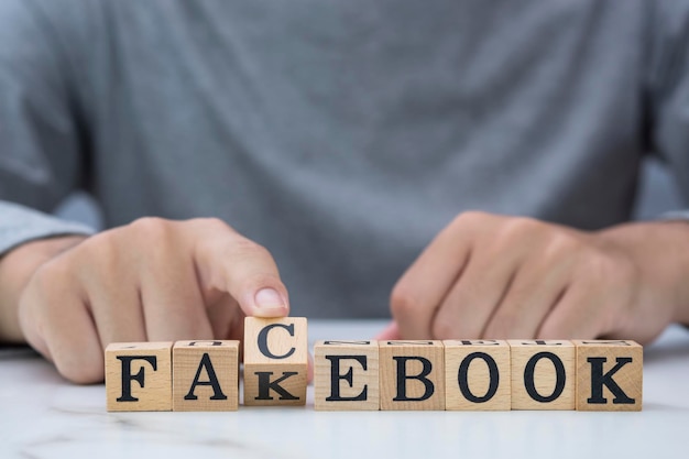 Junger Mann dreht mit der Hand kubische Holzblöcke um, um Fakebook in Facebook-Wortlaut zu ändern