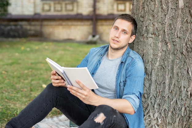 Junger Mann entspannt sich und liest Notizbuch, während er auf grünem Rasen sitzt und aus der Ferne im Park lernt