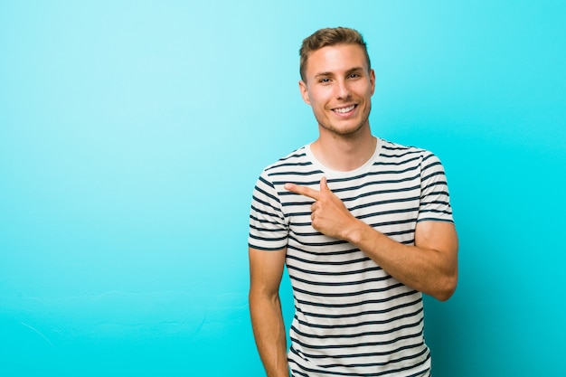 Junger Mann gegen eine blaue Wand, die beiseite lächelt und zeigt und etwas auf Leerstelle zeigt
