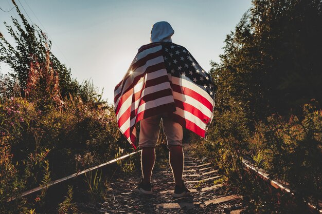 Junger Mann geht mit einer amerikanischen Flagge in Richtung Sonnenuntergang entlang der Bahngleise der Straße. Männlich steht auf einer verlassenen Straße mit Schienen im ländlichen Gebiet mit Streifen und Sternenflagge. Konzept der Reise, Freiheit