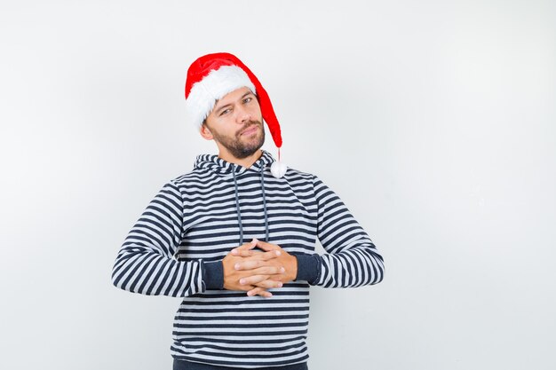 Junger Mann hält die Hände in Hoodie, Weihnachtsmütze und sieht selbstbewusst aus,